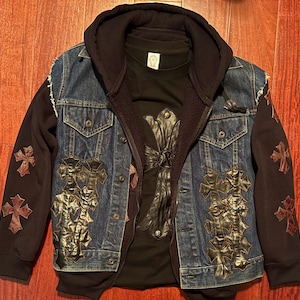 Può includere: Giacca di jeans con cappuccio nero attaccato, con applicazioni di croci in pelle e disegni di croci sulle maniche. Sotto, una maglietta nera con un grande disegno a croce.