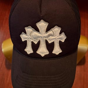 Puede incluir: Gorra de camionero negra con parte trasera de malla y visera curva. La parte delantera presenta un emblema blanco cosido de tres cruces conectadas. Un pequeño emblema plateado está en la parte superior.
