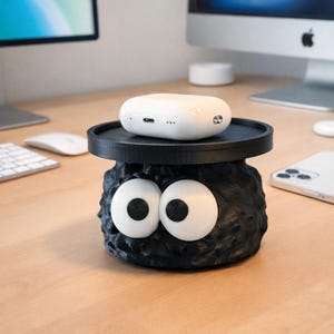 May include: A whimsical black and white decorative item resembling a soot sprite, with large, expressive eyes. A white, oval-shaped electronic device rests on a black circular platform atop the sprite. The item is on a wooden desk.