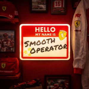May include: A red and white illuminated sign with the text "HELLO MY NAME IS SMOOTH OPERATOR" in a playful font. The sign features the Ferrari logo and star accents, set against a red wall with Ferrari memorabilia.