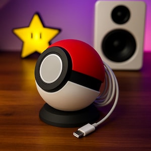 Op de afbeelding: Een rode, witte en zwarte Poké Ball-vormige luidspreker met een zilveren luidspreker en een witte oplaadkabel. De luidspreker staat op een zwarte basis. Een geel stervormig licht staat op de achtergrond.