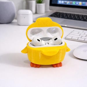 Puede incluir: Una funda amarilla con forma de pato que contiene auriculares inalámbricos blancos. La funda tiene patas naranjas y pequeñas alas. Los auriculares están en una funda abierta sobre un escritorio blanco, con un portátil y otros accesorios en el fondo.