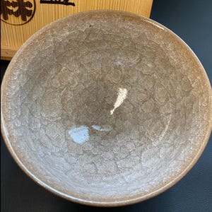 May include: A round ceramic bowl featuring a glossy, light brown glaze with a distinct, textured crackle pattern across the interior surface. The bowl has a smooth, curved rim and is positioned in front of a light-colored wooden box with dark text.