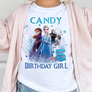 May include: A young child wearing a white t-shirt with the text "CANDY BIRTHDAY GIRL" and a graphic of characters from the movie Frozen. The child is also wearing a pink cardigan, a pink headband with a bow, and round sunglasses.