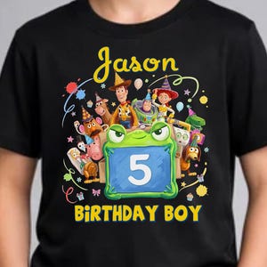 Pu&ograve; includere: T-shirt nera con grafica colorata con la scritta "Jason" e "Birthday Boy". La grafica include personaggi dei cartoni animati e il numero 5 su sfondo blu. La maglietta &egrave; indossata da una persona.