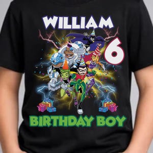 May include: Black t-shirt featuring a colorful graphic of cartoon superheroes with the name "WILLIAM" and the number "6". The text "BIRTHDAY BOY" is printed below the graphic. The shirt is worn by a child.