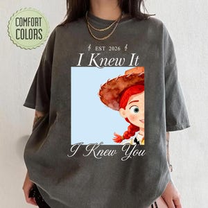 May include: A charcoal gray short-sleeve t-shirt featuring a graphic of a red-haired character wearing a brown cowboy hat. The text reads "EST 2026", "I Knew It", and "I Knew You". A circular "COMFORT COLORS" logo is visible in the top left corner.