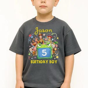 Pu&ograve; includere: Una maglietta grigio scuro a maniche corte con una grafica colorata di personaggi dei cartoni animati e la scritta "Jason" e "Birthday Boy". Il numero "5" &egrave; visualizzato su un cartello blu. La maglietta &egrave; indossata da un bambino.