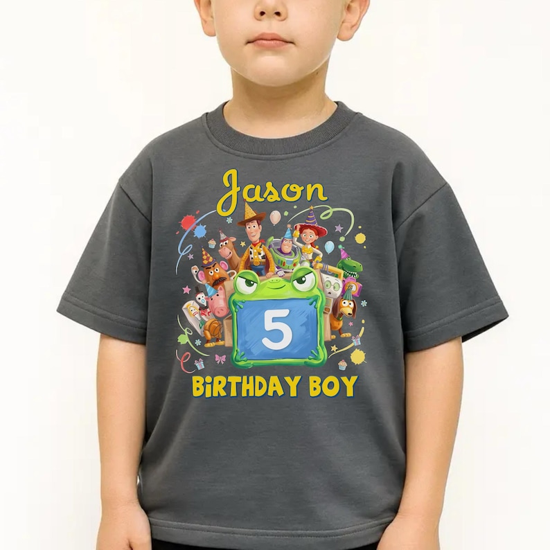 Pu&ograve; includere: Una maglietta grigio scuro a maniche corte con una grafica colorata di personaggi dei cartoni animati e la scritta "Jason" e "Birthday Boy". Il numero "5" &egrave; visualizzato su un cartello blu. La maglietta &egrave; indossata da un bambino.