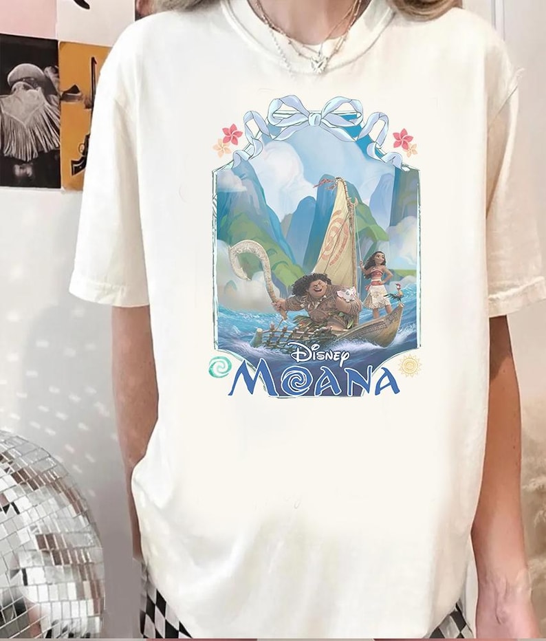 Puede incluir: Una camiseta de manga corta de color crema con un estampado gr&aacute;fico de los personajes de la pel&iacute;cula Disney Moana, Maui y Moana, en un barco. El dise&ntilde;o incluye una cinta azul en la parte superior, flores tropicales y el logo Disney Moana.