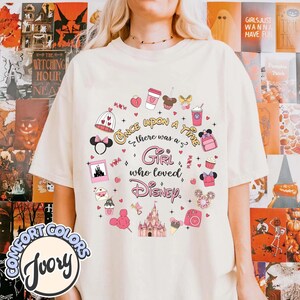 Puede incluir: Camiseta de color marfil con un dise&ntilde;o circular con ilustraciones tem&aacute;ticas de Disney y el texto "Once upon a time there was a Girl who loved Disney." El dise&ntilde;o incluye un castillo, orejas de Mickey Mouse y otros iconos.