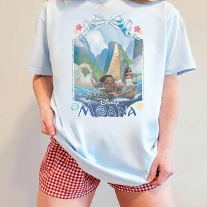Puede incluir: Una camiseta azul claro con un estampado gr&aacute;fico de los personajes de Disney Moana, Maui y Moana, en un barco. El dise&ntilde;o incluye una cinta azul claro en la parte superior, flores tropicales y el texto Disney Moana en letras azules abajo.