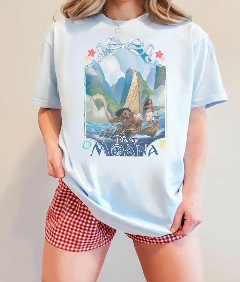 Puede incluir: Una camiseta azul claro con un estampado gr&aacute;fico de los personajes de Disney Moana, Maui y Moana, en un barco. El dise&ntilde;o incluye una cinta azul claro en la parte superior, flores tropicales y el texto Disney Moana en letras azules abajo.