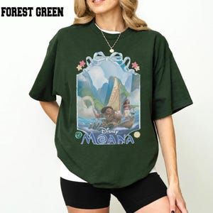 Puede incluir: Una camiseta de manga corta en color verde bosque con un estampado gr&aacute;fico de los personajes de Disney Moana, Maui y Moana, en un barco. El dise&ntilde;o est&aacute; enmarcado por una cinta azul claro e incluye el texto Disney Moana en azul.