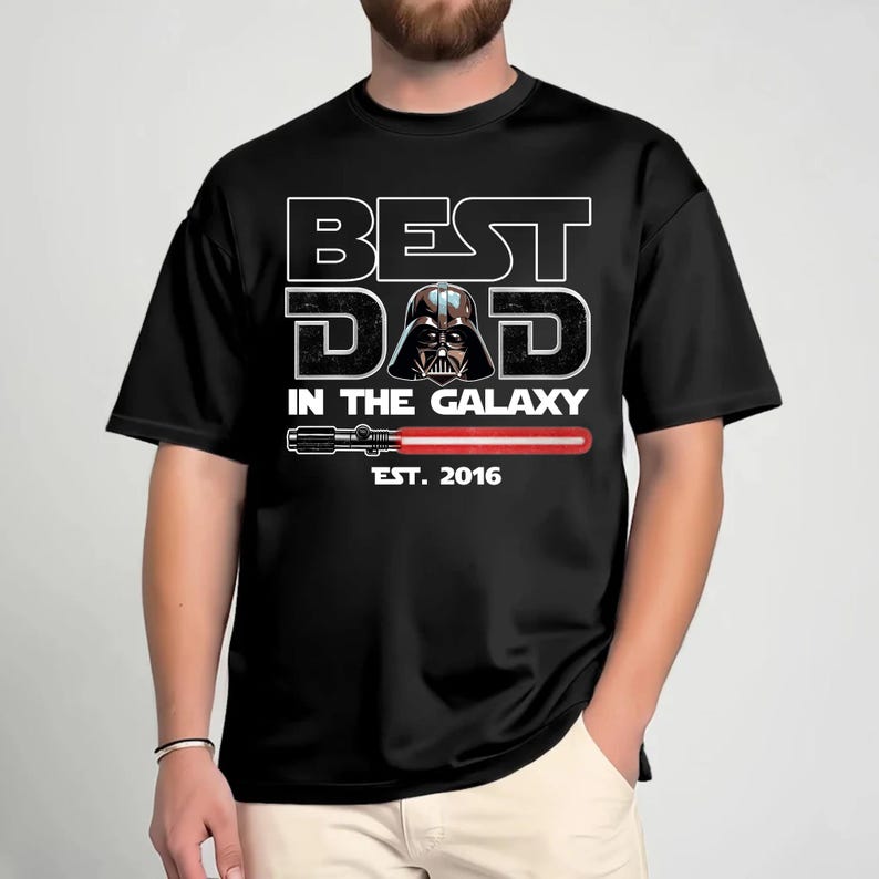 K&ouml;nnte beinhalten: Ein schwarzes T-Shirt mit einem Aufdruck, der den Text BEST DAD IN THE GALAXY zeigt. Das Design enth&auml;lt einen Darth-Vader-Helm, der den Buchstaben A ersetzt, eine rote Lichtschwert-Grafik und den Text EST. 2016 in wei&szlig;en Buchstaben.