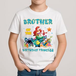 May include: White t-shirt with a colorful graphic featuring Ariel from The Little Mermaid, a lobster, and other sea creatures. Text reads "Brother of the Birthday Princess" in blue and yellow.
