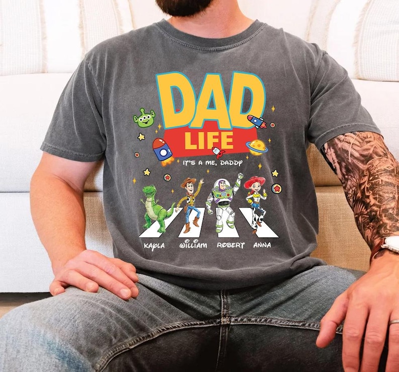 May include: Gray t-shirt with the words "DAD LIFE" in yellow and orange, featuring cartoon characters and the text "IT'S A ME, DADDY." Below the characters are names. The shirt has a vintage, worn look.