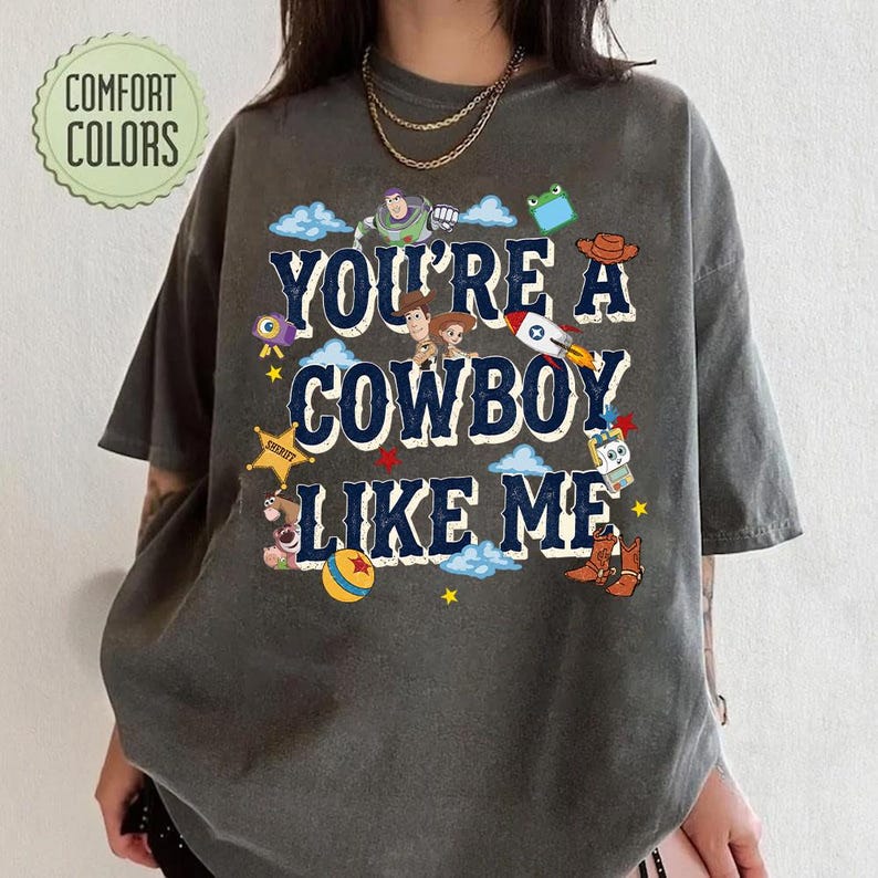May include: A charcoal gray short-sleeve t-shirt featuring the text "YOU'RE A COWBOY LIKE ME" in dark blue letters. The design includes various cartoon characters, a rocket, a yellow star, a ball, and cowboy boots scattered around the text.