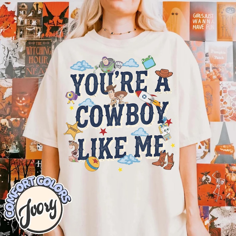 May include: An ivory t-shirt featuring the dark blue text "YOU'RE A COWBOY LIKE ME" surrounded by colorful illustrations of characters and icons from the Toy Story franchise, including a space ranger, a cowboy, a rocket, a ball, and boots.