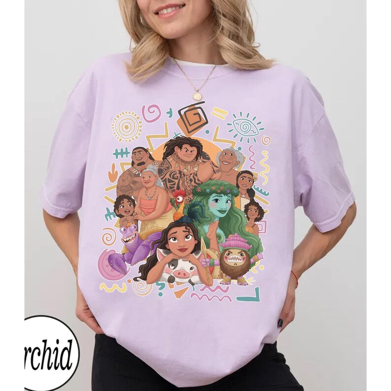 May include: A light purple short-sleeve t-shirt featuring a colorful graphic print of various animated characters from the movie Moana, including Moana, Maui, Pua, Heihei, and Tamatoa, surrounded by stylized geometric shapes and tribal patterns.