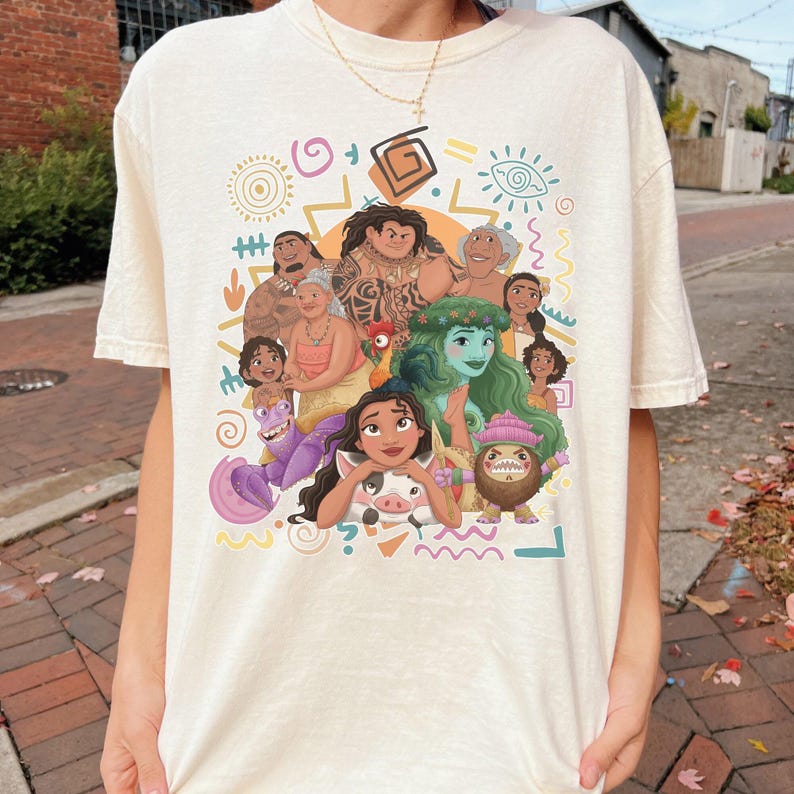 May include: A cream-colored t-shirt featuring a colorful graphic print of various animated characters from the movie Moana, including Moana, Maui, Pua, Heihei, and Tamatoa, surrounded by abstract yellow, pink, and teal geometric shapes and symbols.