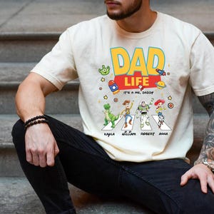 May include: Cream-colored t-shirt with a colorful "DAD LIFE" graphic. The design includes cartoon characters crossing a street, with names below. The shirt also features rockets, stars, and an alien.