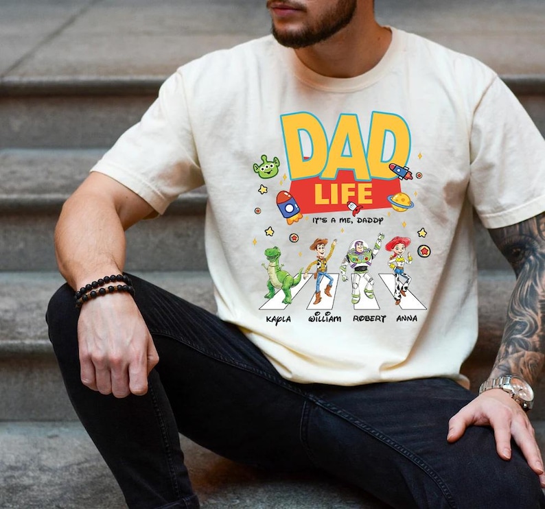 May include: Cream-colored t-shirt with a colorful "DAD LIFE" graphic. The design includes cartoon characters crossing a street, with names below. The shirt also features rockets, stars, and an alien.