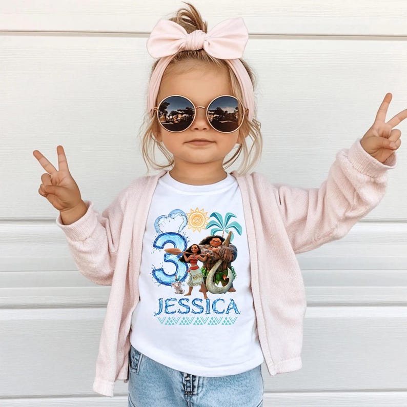 Peut inclure: Un t-shirt blanc avec une illustration de Moana et Maui, un grand chiffre trois bleu, un soleil, des palmiers et le nom JESSICA en bleu. Le t-shirt est port&eacute; avec un gilet rose clair, un bandeau &agrave; n&oelig;ud rose assorti et des lunettes rondes.