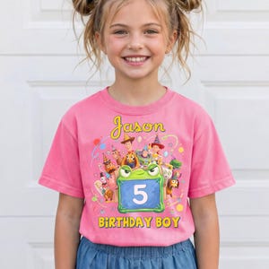 Pu&ograve; includere: T-shirt rosa a maniche corte con una grafica colorata con personaggi dei cartoni animati e le scritte "Jason" e "Birthday Boy". Un personaggio verde a forma di rana mostra il numero "5" sulla pancia.