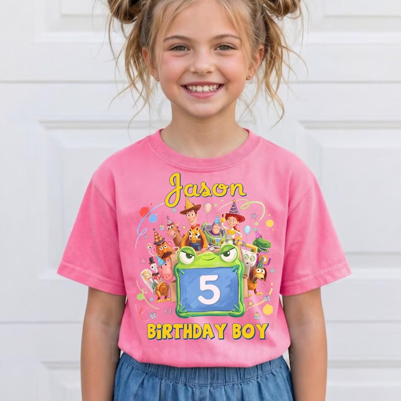 Pu&ograve; includere: T-shirt rosa a maniche corte con una grafica colorata con personaggi dei cartoni animati e le scritte "Jason" e "Birthday Boy". Un personaggio verde a forma di rana mostra il numero "5" sulla pancia.