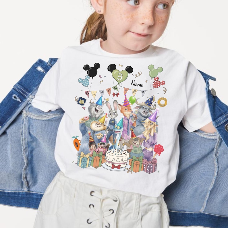 Pu&ograve; includere: T-shirt bianca con una grafica colorata di personaggi dei cartoni animati, una torta di compleanno e la scritta "Name". Il design include palloncini e il numero "3". La maglietta &egrave; indossata da una bambina.