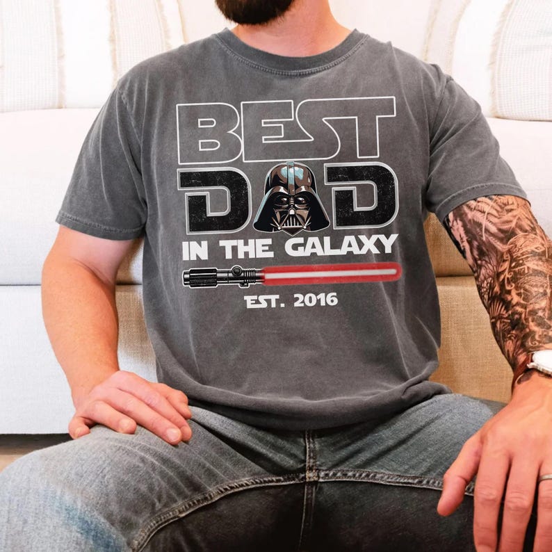 K&ouml;nnte beinhalten: Ein anthrazitfarbenes Kurzarm-T-Shirt mit einem Grafikdruck, der BEST DAD IN THE GALAXY liest, sowie einer Abbildung von Darth Vaders Helm, einem roten Lichtschwert und dem Text EST. 2016 in wei&szlig;er und schwarzer Schrift.