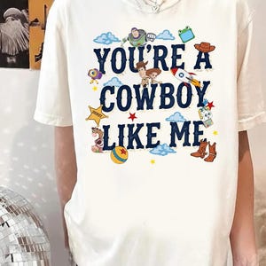 May include: A cream-colored short-sleeve t-shirt featuring the dark blue text "YOU'RE A COWBOY LIKE ME" surrounded by colorful cartoon graphics of characters, a rocket, a sheriff star, a ball, and cowboy boots in a playful, scattered arrangement.