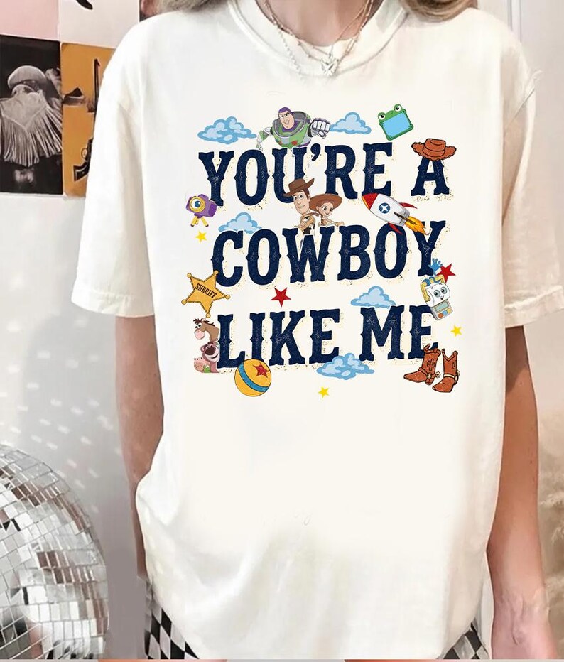 May include: A cream-colored short-sleeve t-shirt featuring the dark blue text "YOU'RE A COWBOY LIKE ME" surrounded by colorful cartoon graphics of characters, a rocket, a sheriff star, a ball, and cowboy boots in a playful, scattered arrangement.