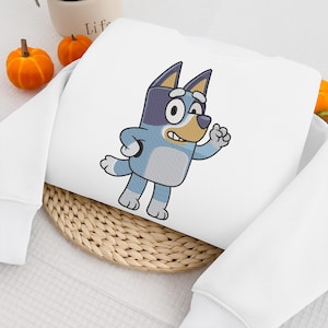 Peut inclure: Sweat-shirt blanc avec un personnage de dessin animé de chien brodé en bleu, gris et beige. Le chien est représenté en train de courir. Le sweat-shirt est posé sur un tapis tissé, avec de petites citrouilles oranges et un pot blanc en arrière-plan.