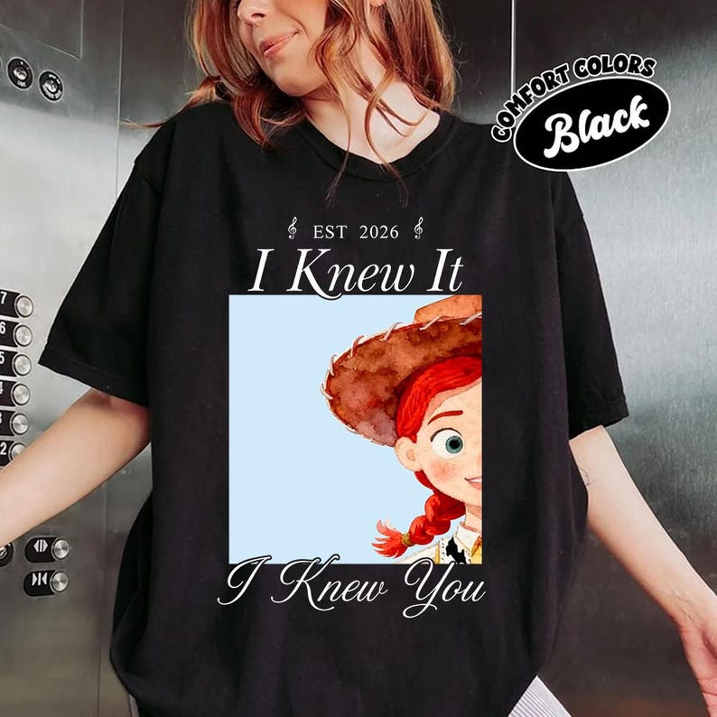 May include: A black short-sleeve t-shirt featuring a graphic print of a cartoon cowgirl with red hair and a brown hat. The text reads EST 2026, I Knew It, and I Knew You. A small oval badge in the top right corner displays the text COMFORT COLORS Black.