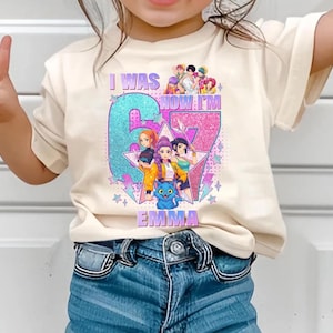 May include: A cream-colored t-shirt with a colorful graphic design. The design features the text "I WAS" above a large number "67" with the name "EMMA" below. The number is decorated with glitter and cartoon characters.