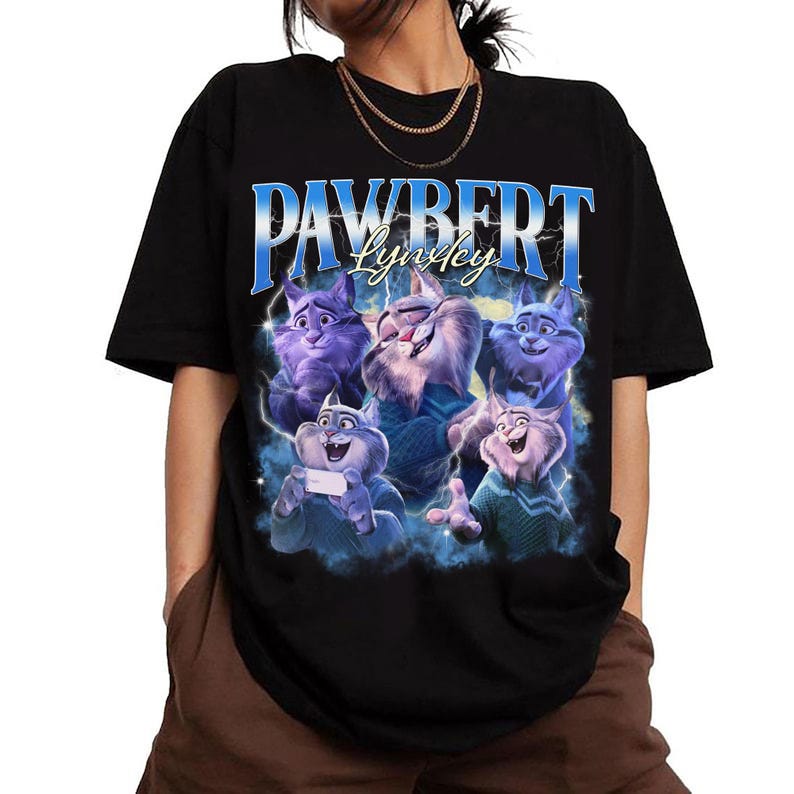 Op de afbeelding: Zwart T-shirt met een afbeelding van vijf cartoonkatten in blauwe en paarse tinten. De tekst "PAWBERT Lynley" is in blauw en wit gedrukt. De katten staan tegen een blauw-witte, bewolkte achtergrond.