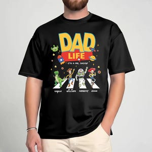 May include: Black t-shirt with a colorful graphic featuring the words "DAD LIFE" in yellow and blue, with the phrase "IT'S ME, DADDY" below. The design includes cartoon characters crossing a street, with names below them.