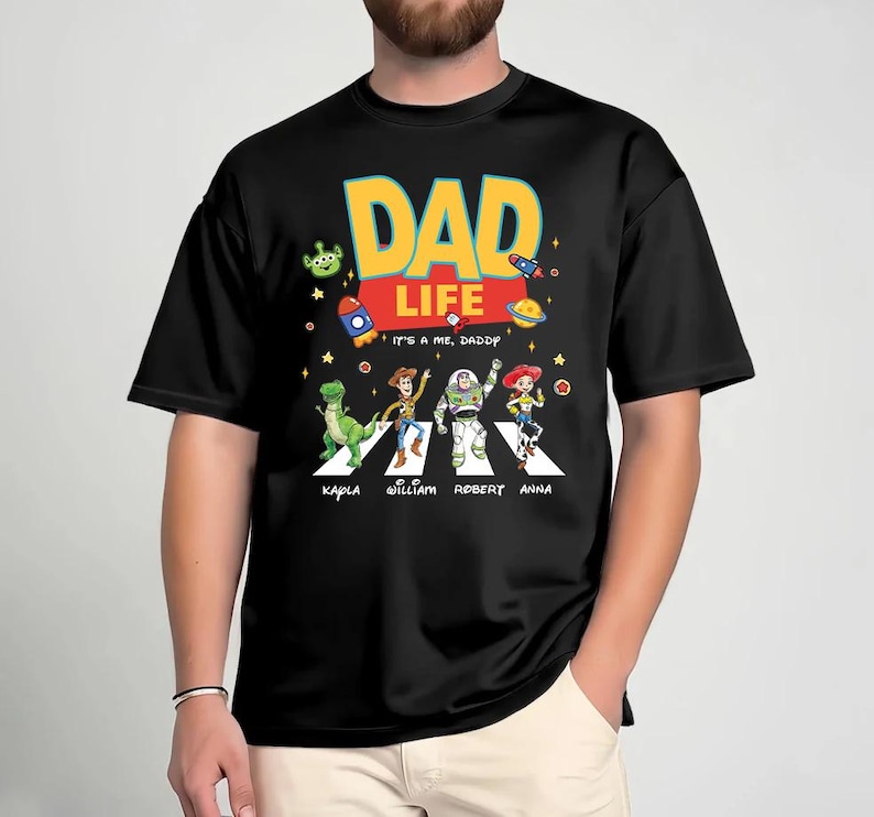 May include: Black t-shirt with a colorful graphic featuring the words "DAD LIFE" in yellow and blue, with the phrase "IT'S ME, DADDY" below. The design includes cartoon characters crossing a street, with names below them.