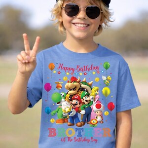 May include: A blue t-shirt featuring a colorful graphic of video game characters, balloons, and the text "Happy Birthday." Below the graphic, the shirt reads "Brother Of The Birthday Boy." The person in the image is wearing a blue baseball cap and sunglasses.