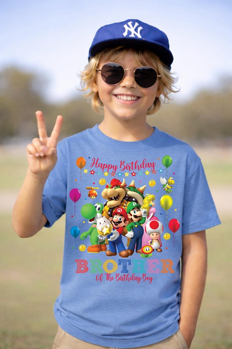 May include: A blue t-shirt featuring a colorful graphic of video game characters, balloons, and the text "Happy Birthday." Below the graphic, the shirt reads "Brother Of The Birthday Boy." The person in the image is wearing a blue baseball cap and sunglasses.