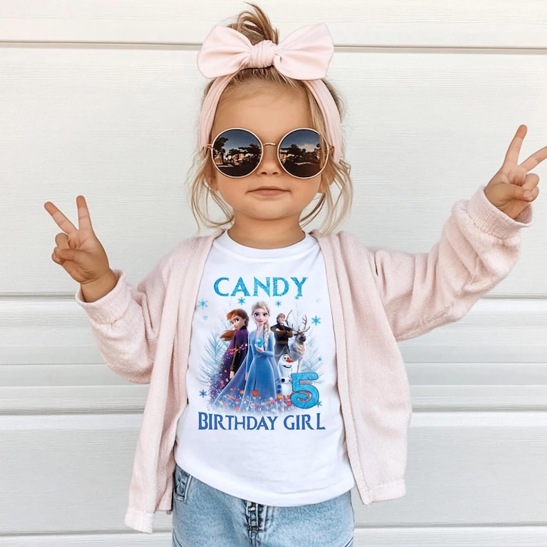 May include: A young child wearing a white t-shirt with the text "CANDY BIRTHDAY GIRL" and a graphic of characters from the movie Frozen. The child is also wearing a pink cardigan, a pink headband with a bow, and round sunglasses.
