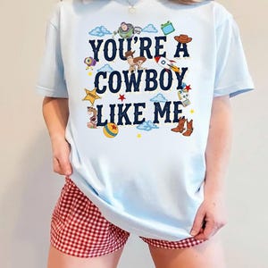 May include: A light blue t-shirt featuring the dark blue text "YOU'RE A COWBOY LIKE ME" surrounded by various colorful cartoon character illustrations, including a space ranger, a sheriff, a cowgirl, a rocket, a ball, and cowboy boots.