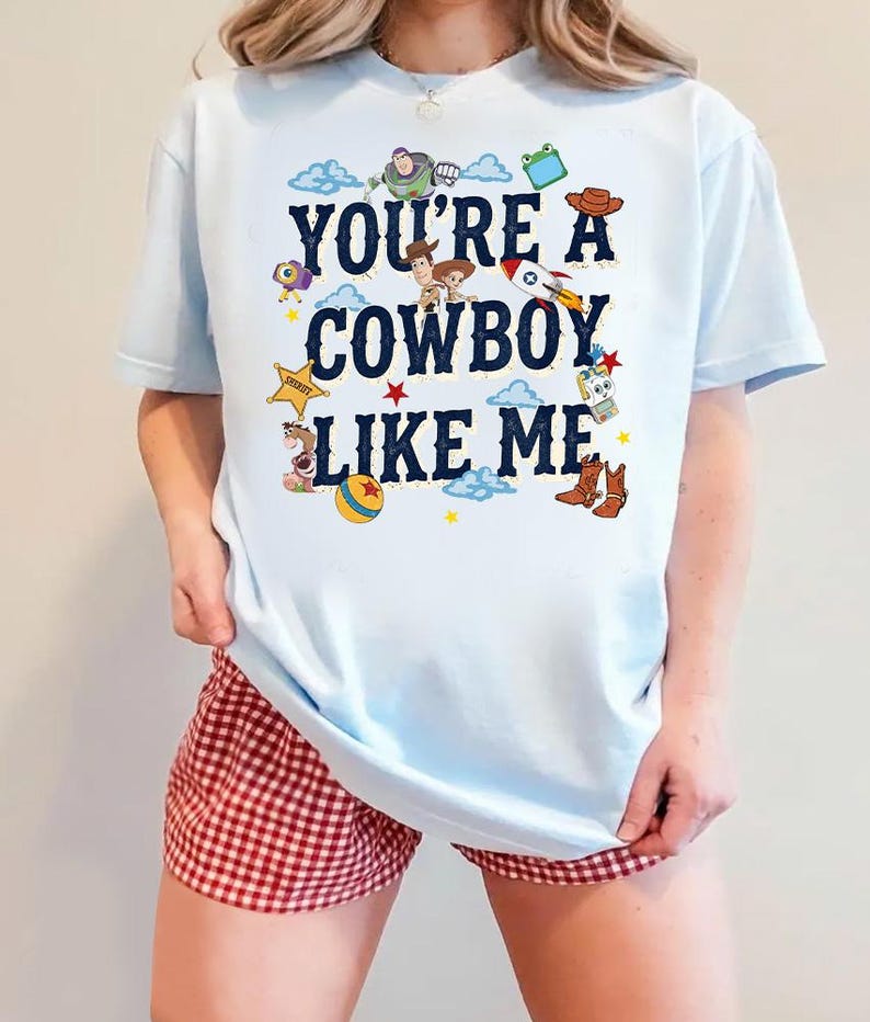 May include: A light blue t-shirt featuring the dark blue text "YOU'RE A COWBOY LIKE ME" surrounded by various colorful cartoon character illustrations, including a space ranger, a sheriff, a cowgirl, a rocket, a ball, and cowboy boots.