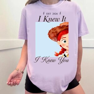 May include: A light purple t-shirt featuring a graphic print of a cartoon character with red hair and a brown hat. The text "EST 2026", "I Knew It", and "I Knew You" is printed in black above and below the square image of the character.