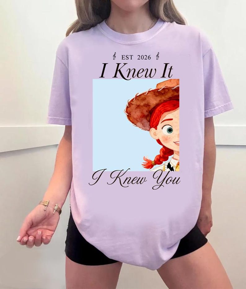 May include: A light purple t-shirt featuring a graphic print of a cartoon character with red hair and a brown hat. The text "EST 2026", "I Knew It", and "I Knew You" is printed in black above and below the square image of the character.