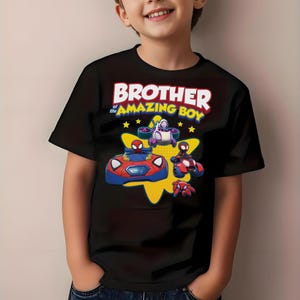 May include: A black short-sleeved t-shirt featuring a graphic print with the text BROTHER of the AMAZING BOY in white and yellow. The design includes cartoon characters in red and blue vehicles against a yellow star shape with small yellow stars.