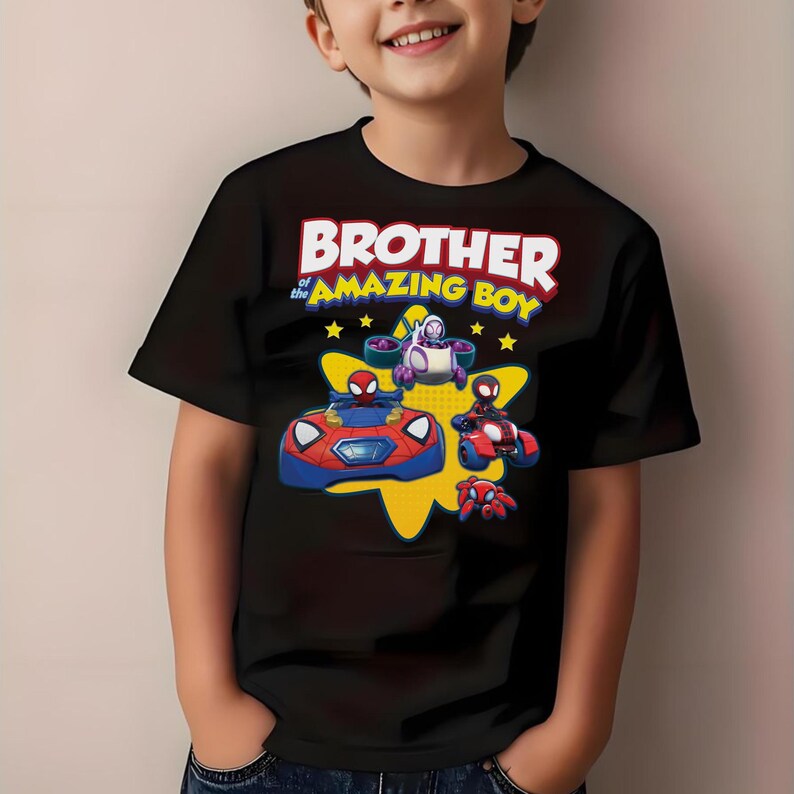 May include: A black short-sleeved t-shirt featuring a graphic print with the text BROTHER of the AMAZING BOY in white and yellow. The design includes cartoon characters in red and blue vehicles against a yellow star shape with small yellow stars.