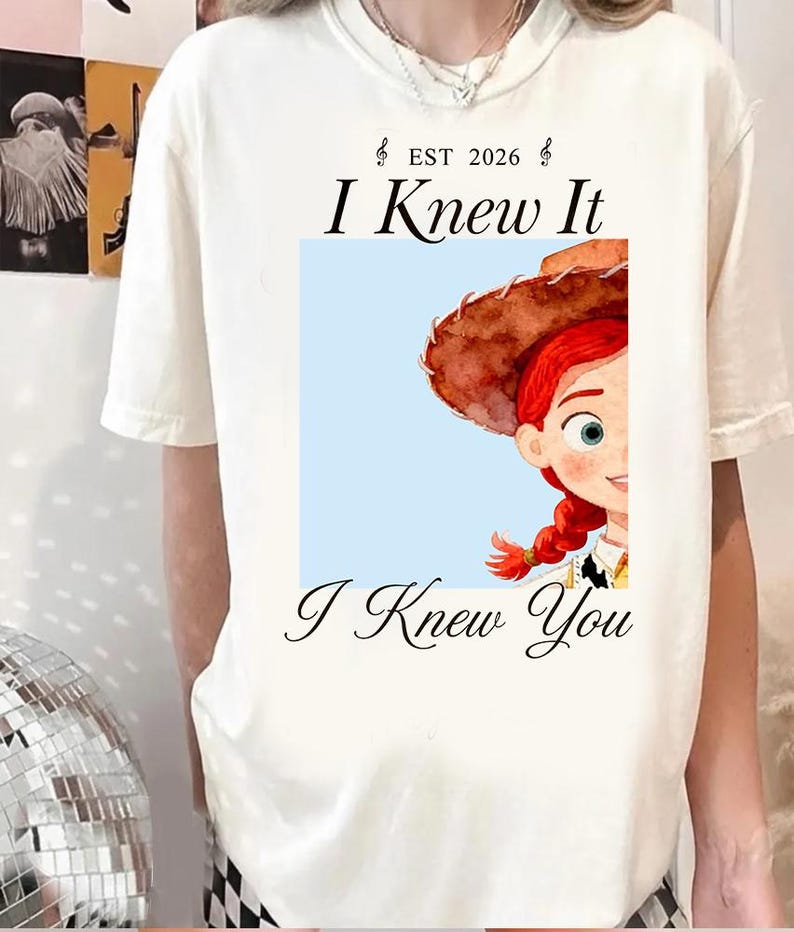 May include: A white short-sleeve t-shirt featuring a graphic of a red-haired character wearing a brown cowboy hat. The text "EST 2026", "I Knew It", and "I Knew You" is printed in black above and below the square illustration on the front.